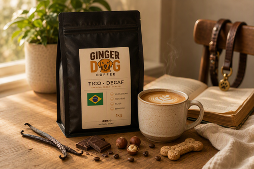 A black bag of Ginger Dog Coffee "Tico Decaf" medium roast coffee stands on a wooden table beside a steaming latte with latte art. Around the coffee are vanilla pods, chocolate pieces, coffee beans, hazelnuts, and a dog-shaped biscuit. In the background, an open book, a dog lead draped over a chair, and a potted plant are softly lit by warm natural sunlight, creating a cosy home setting.